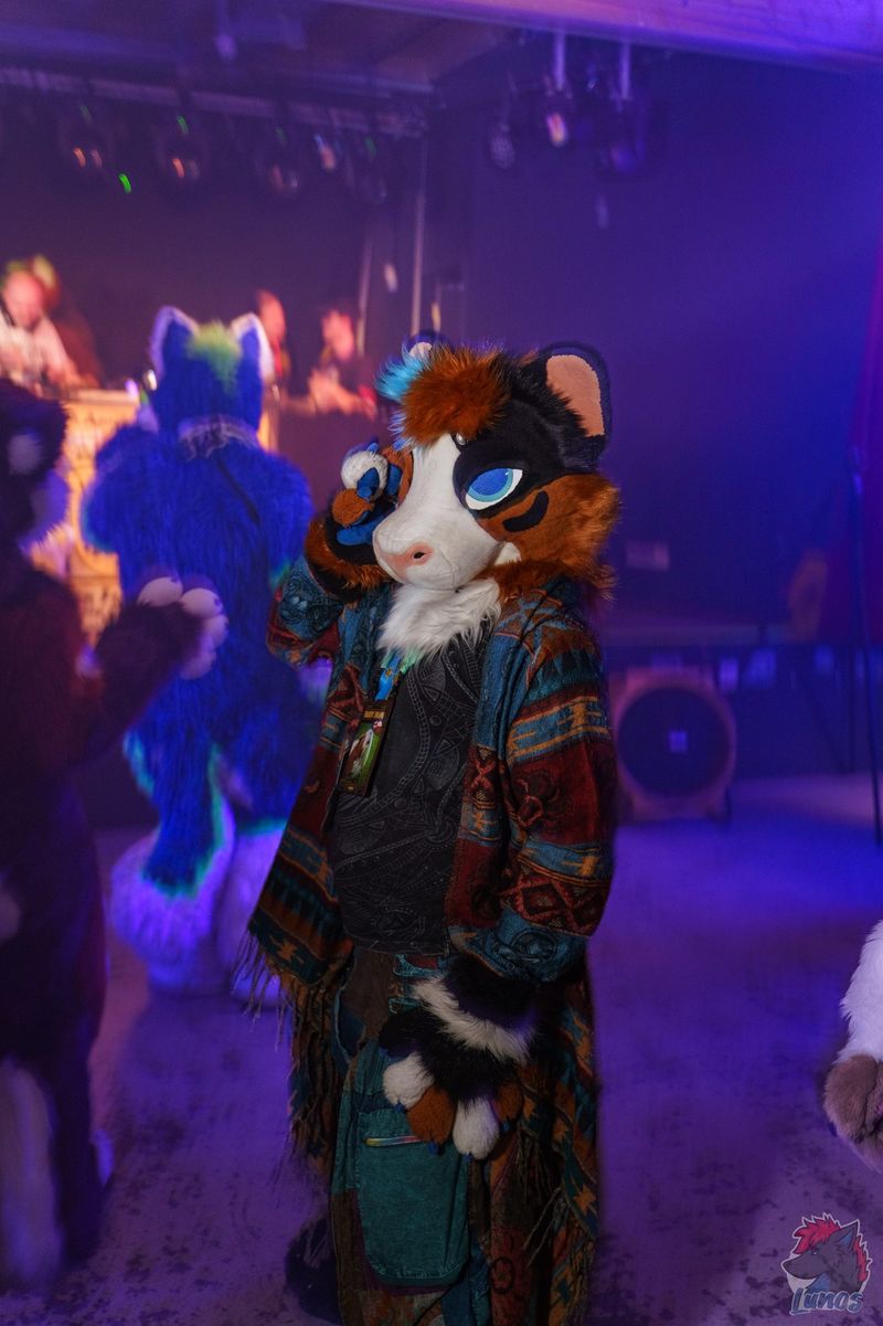 Drapii in fursuit and a patterned fringed cardigan on a purple-lit dance floor, with other fursuiters dancing behind